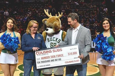 Sherri Tussler, Executive Director of Hunger Task Force, accepts check from Justin McCoy, Vice President of Marketing at Cousins Subs. (Also pictures: Bango, Milwaukee Bucks mascot, and Bucks dancers.)